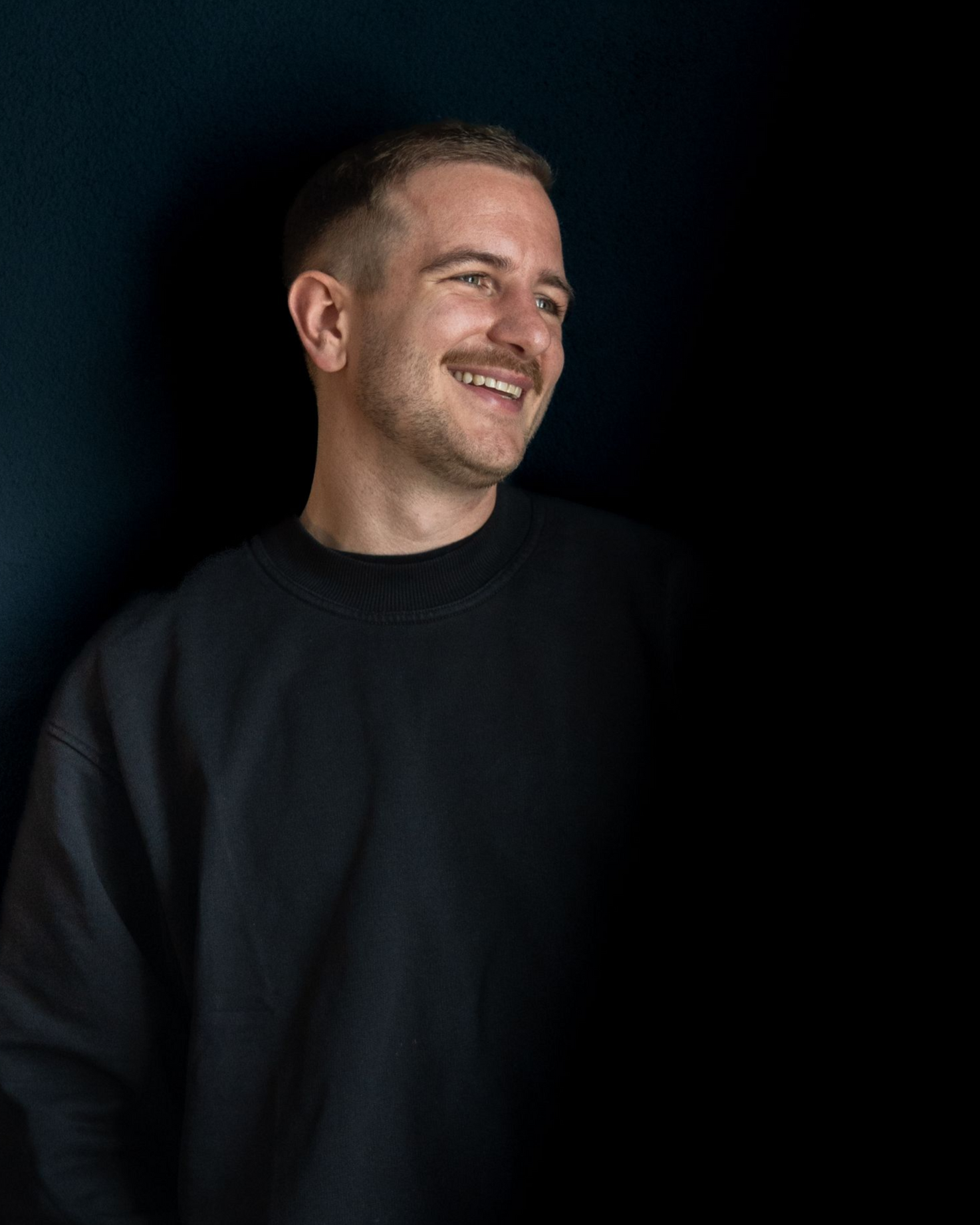 Ein lächelnder Mann in einem schwarzen Pullover vor dunklem Hintergrund blickt zur Seite.
