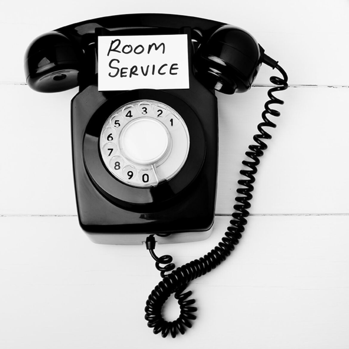 Schwarzes Wählscheibentelefon mit der Aufschrift „Room Service“ auf der Oberseite, abgebildet auf weißem Hintergrund.