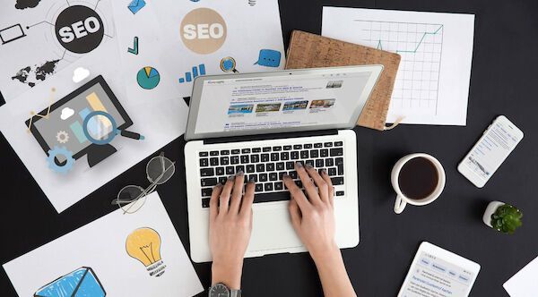 Hände tippen auf einem Laptop mit SEO-Diagrammen, Kaffee, Telefon und Schreibtischutensilien auf einem schwarzen Tisch