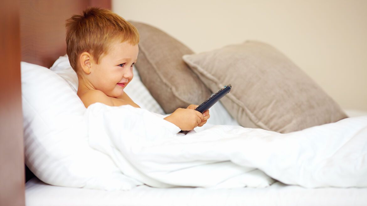 Lächelndes Kind liegt in einem Hotelbett mit einer Fernbedienung in der Hand