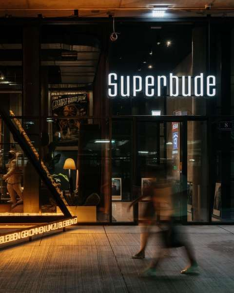 Schaufenster bei Nacht mit leuchtendem „Superbude“-Schild und einem verschwommenen Fußgänger, der vorbeigeht