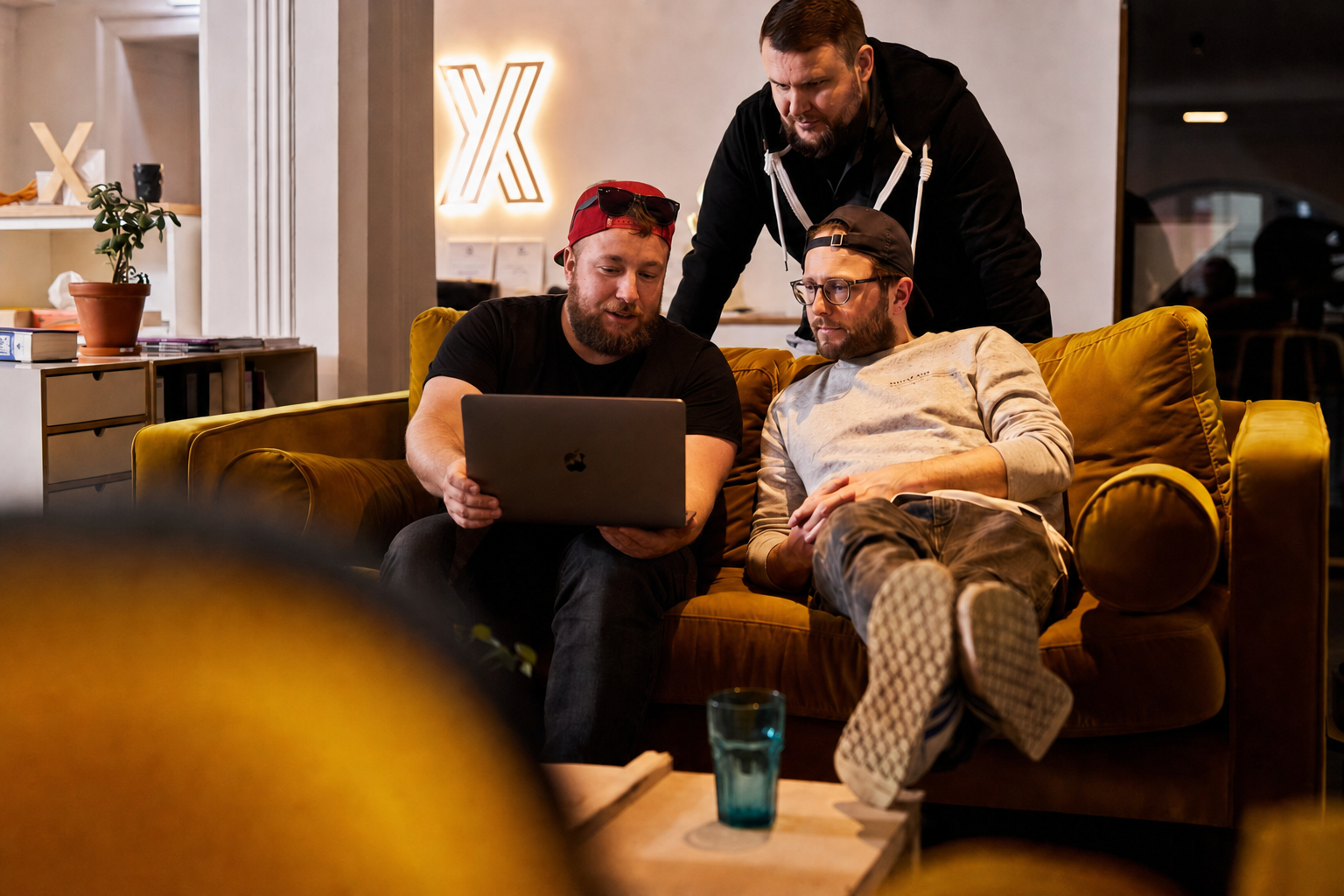 Drei Personen sitzen auf einer Couch und schauen in einem warm beleuchteten Wohnzimmer auf einen Laptop.