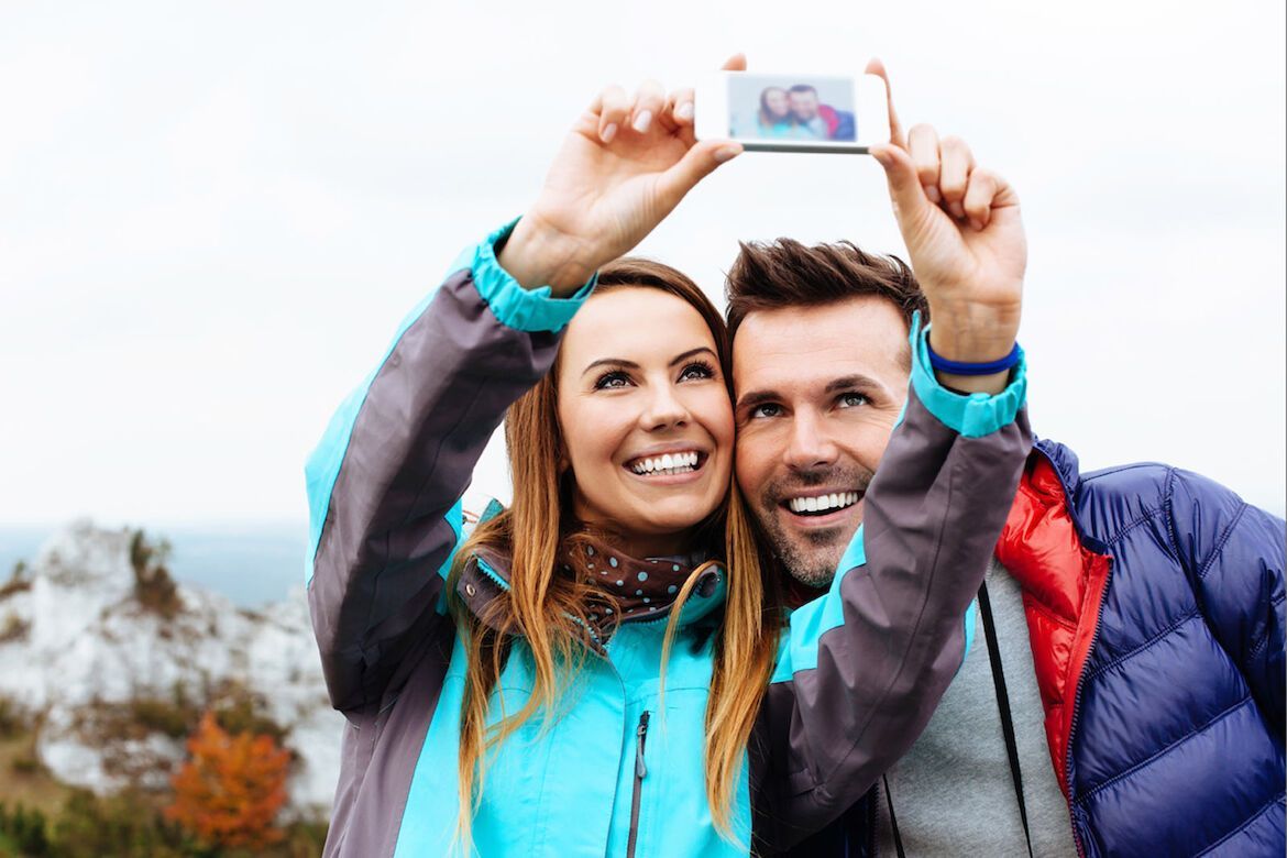 Ein lächelndes Paar macht ein Selfie im Freien, beide tragen blaue Jacken; im Hintergrund sind Herbstbäume zu sehen.