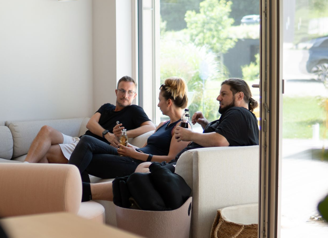 Drei Personen unterhalten sich auf einem Sofa in einem hellen Wohnzimmer mit großen Fenstern.