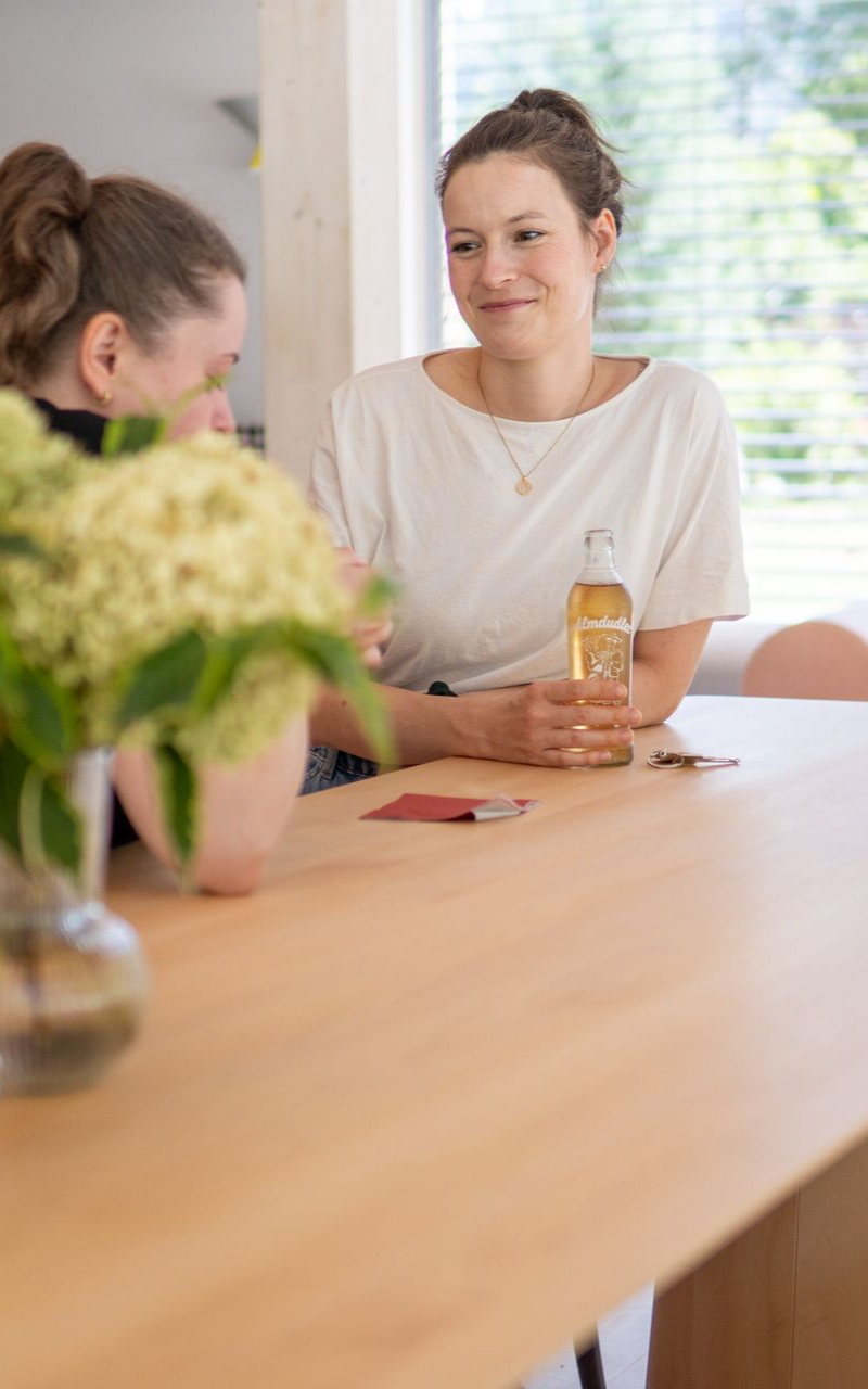 Zwei Frauen unterhalten sich an einem Tisch, eine hält eine Getränkeflasche in der Hand, in einem hellen Café.