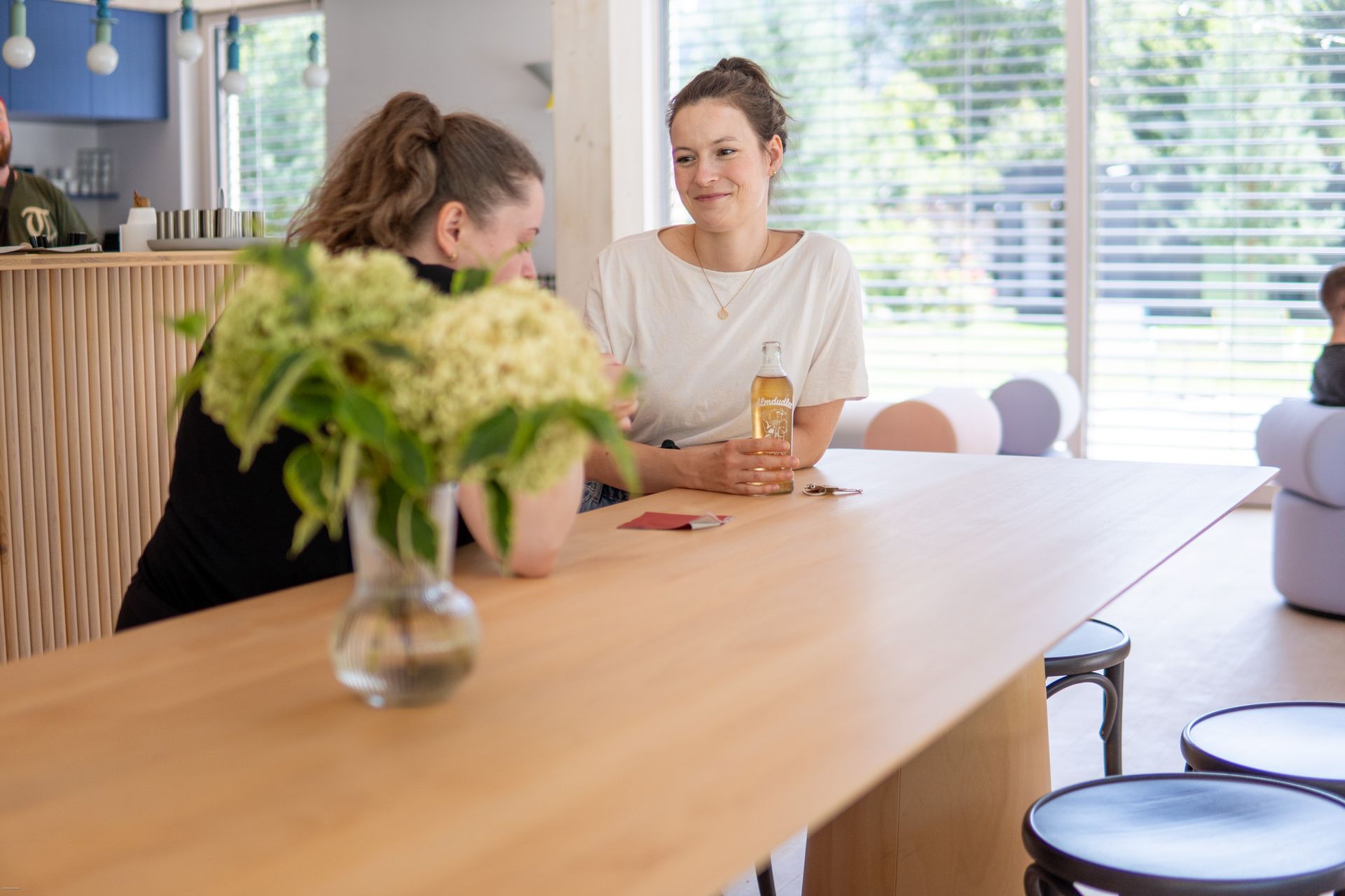 Zwei Personen unterhalten sich an einem Holztisch in einem hellen Café; im Vordergrund steht eine Blumenvase.