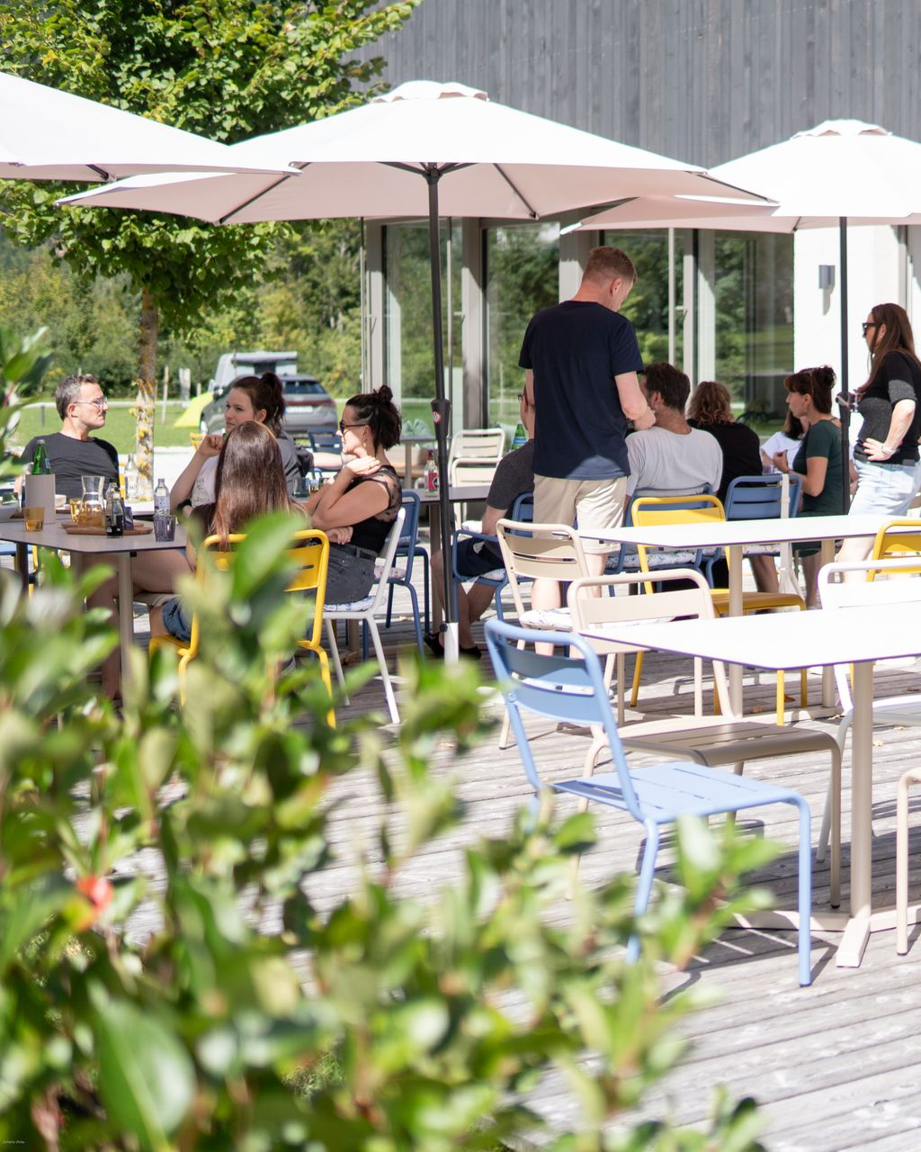 Terrasse eines Cafés im Freien, auf der an einem sonnigen Tag Menschen unter großen Sonnenschirmen speisen.