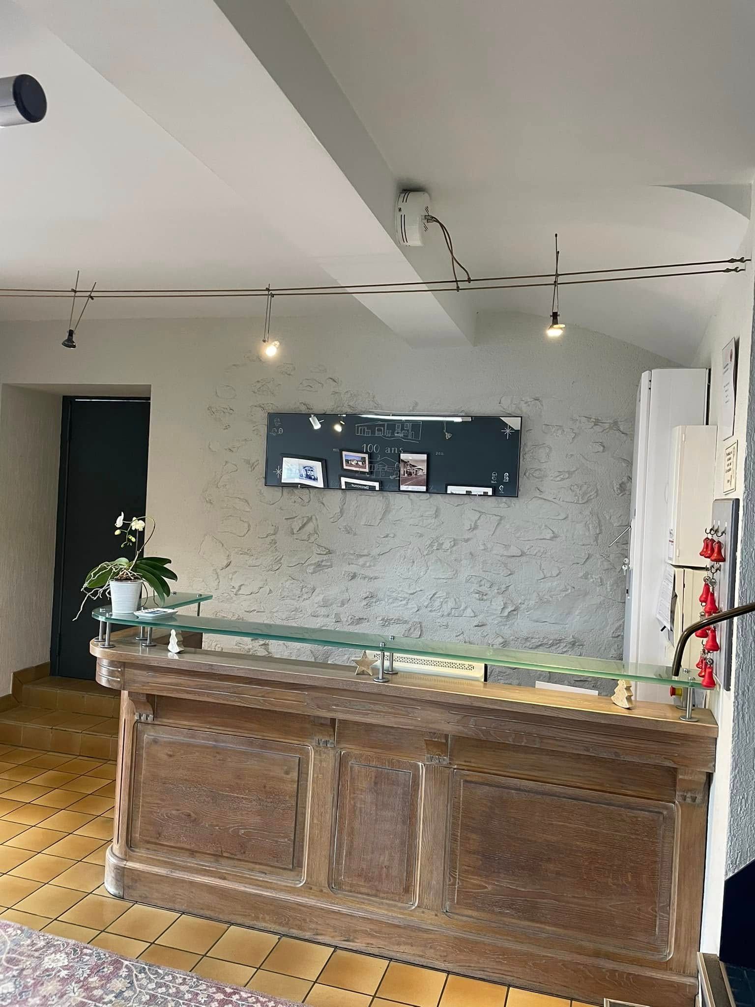 Comptoir d'accueil en bois avec plateau en verre devant un mur blanc texturé. Porte sombre et petites plantes.