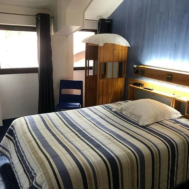 Chambre avec lit, couette à rayures bleues et blanches, chaise bleue, armoire en bois et fenêtres.