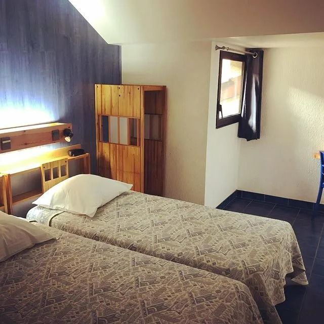 Deux lits jumeaux dans une chambre simple, bureau et armoire en bois, petite fenêtre avec rideau sombre.