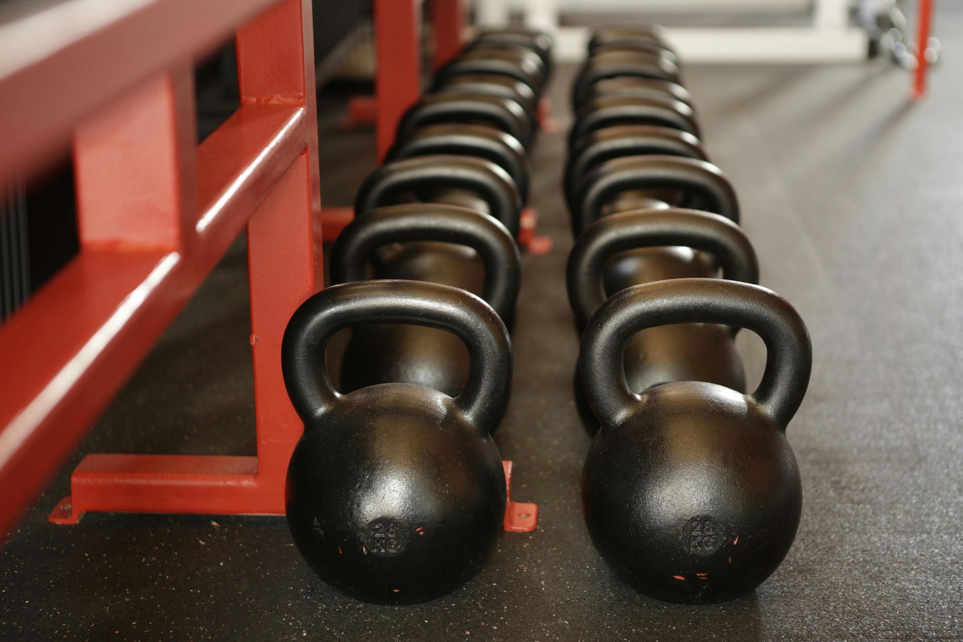 Rows of black kettlebells on a gym floor, lined up near a red metal rack.