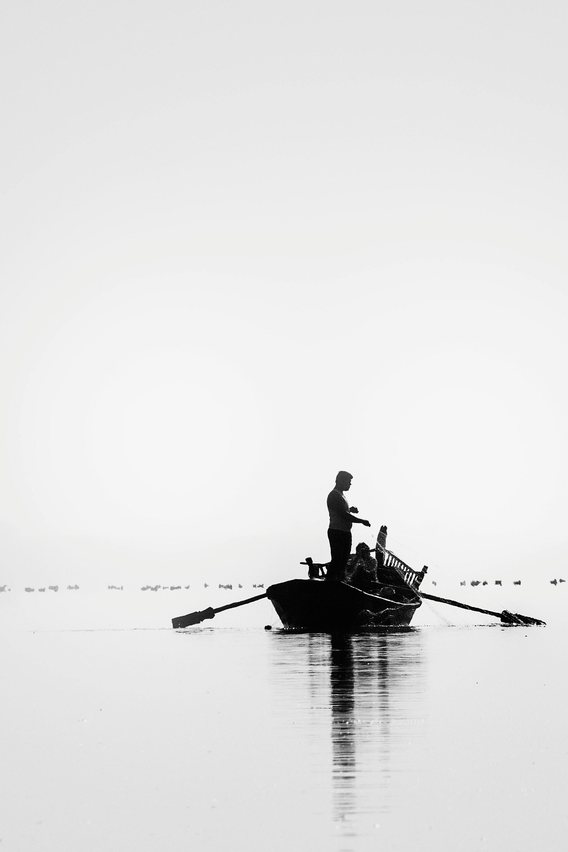 Man rowing boat on calm water. Black and white, silhouetted against a bright sky.