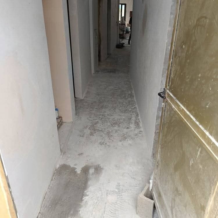 Pasillo estrecho sin terminar con paredes blancas, suelo polvoriento y una puerta parcialmente abierta a la derecha.