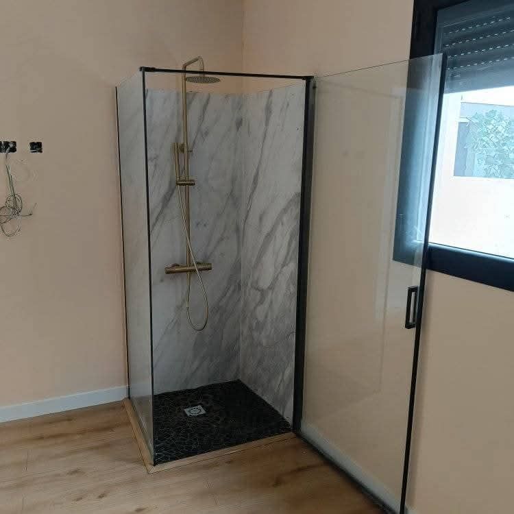 Mampara de ducha de cristal con paneles de pared que imitan el mármol y desagüe oscuro en el suelo en un baño.