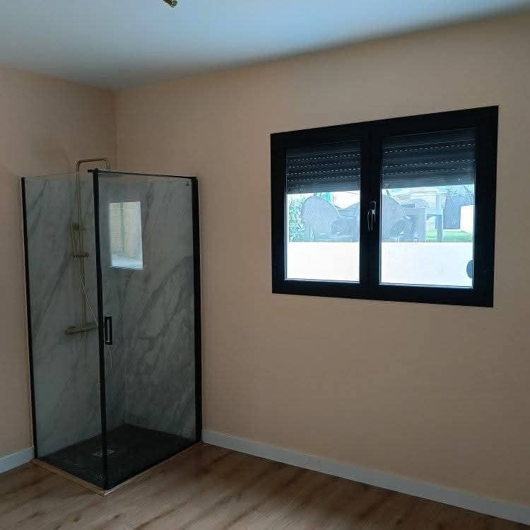 Habitación vacía de color beige con suelo de madera, ventana con marco negro y una cabina de ducha de cristal en la esquina.