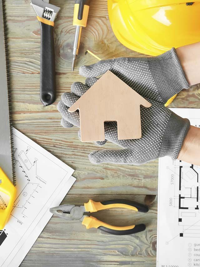 Manos enguantadas sosteniendo una maqueta de una casa de madera con herramientas, planos y un casco amarillo sobre un banco de trabajo.