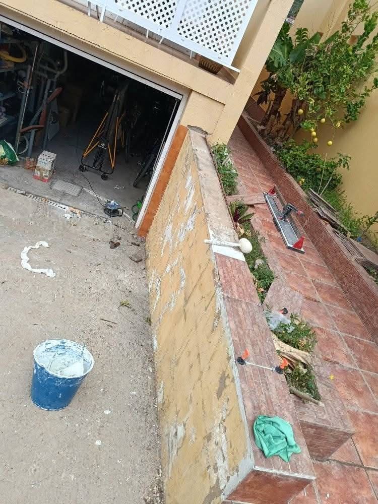 Vista inclinada de la entrada de un edificio con un cubo azul, herramientas y una pared de ladrillos junto a un pequeño jardín.