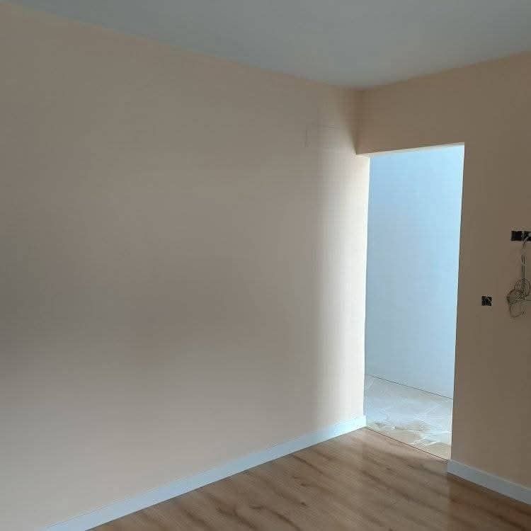 Habitación vacía de color beige con suelo de madera y una puerta abierta que da a un pasillo luminoso.