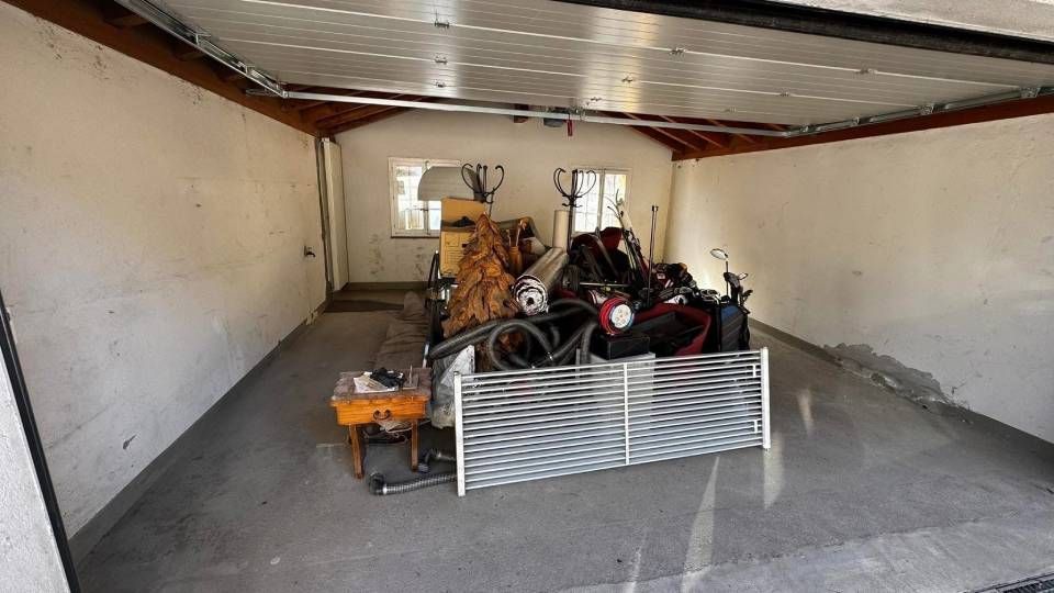 Un garage encombré, aux murs blancs, au sol en béton gris et avec un panneau de portail métallique devant divers objets entreposés.