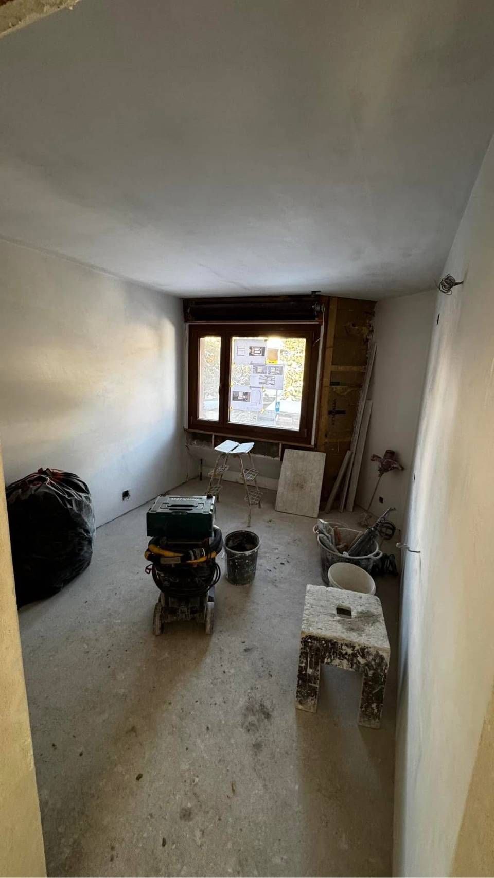 Une pièce en rénovation avec un sol en béton brut, des murs peints en blanc, une fenêtre, des outils de construction et un petit tabouret.