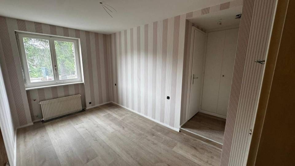 Une pièce vide et non meublée, avec du papier peint à rayures claires, un sol imitation bois, une fenêtre et une porte blanche.