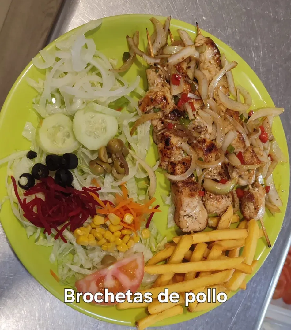Plato de brochetas de pollo con patatas fritas, lechuga, cebolla, pepino, aceitunas y verduras en rodajas.