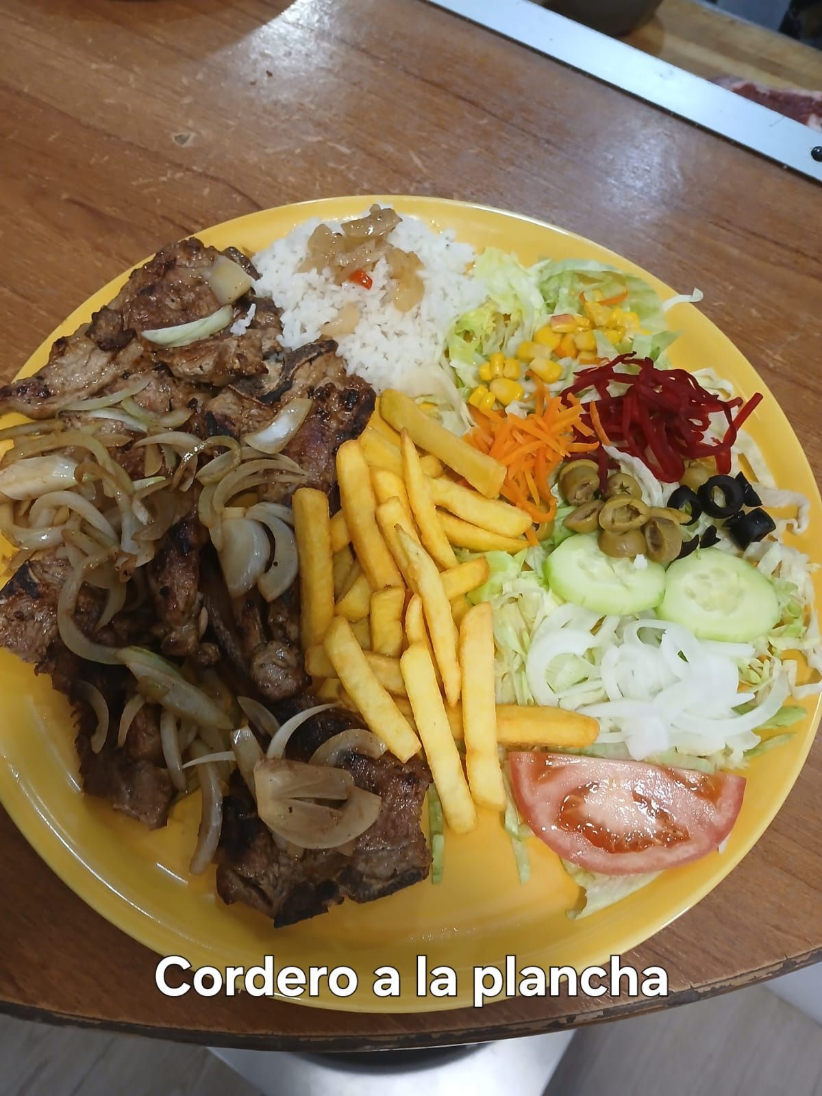Plato de cordero a la plancha con papas fritas, arroz, ensalada y verduras sobre mesa de madera