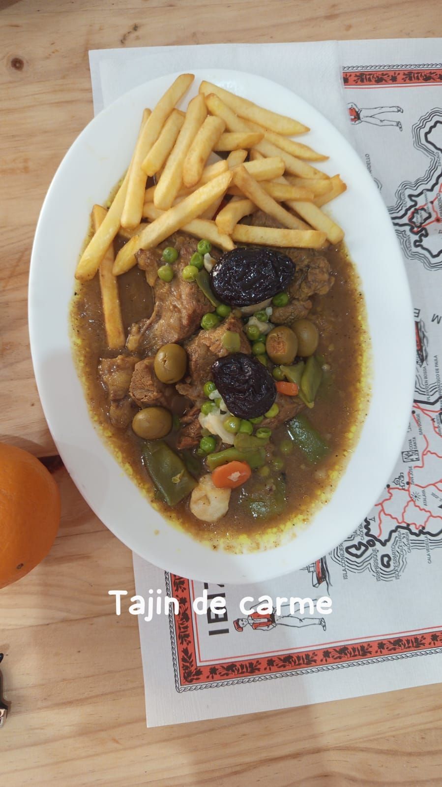 Plato de tajine de carne con patatas fritas y aceitunas verdes sobre un mantel individual estampado, en una mesa de madera.