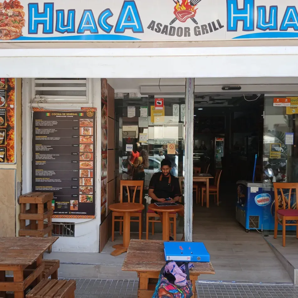 Street view of Huaca Asador Grill with open entrance, menu board, and wooden tables outside