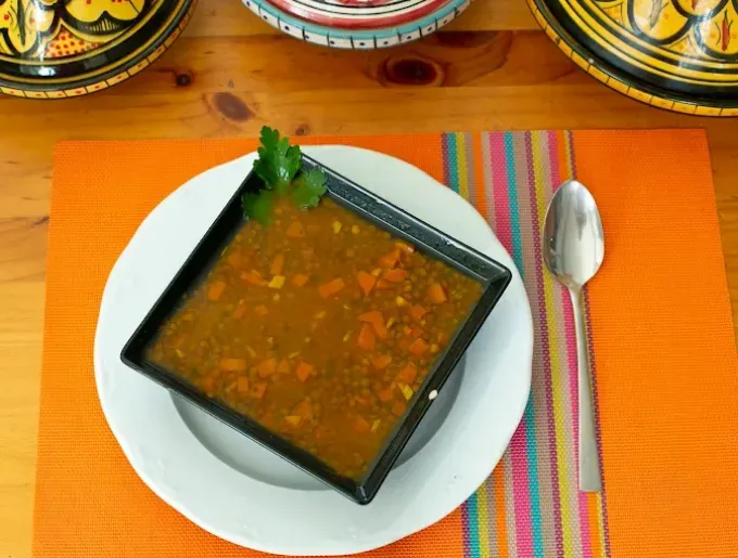 Un tazón negro de sopa sobre un plato blanco con una cuchara sobre un mantel individual naranja.