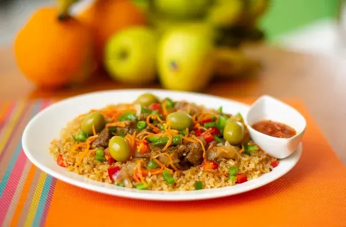 Plato de arroz con verduras y aceitunas sobre un mantel naranja, con fruta difuminada al fondo.