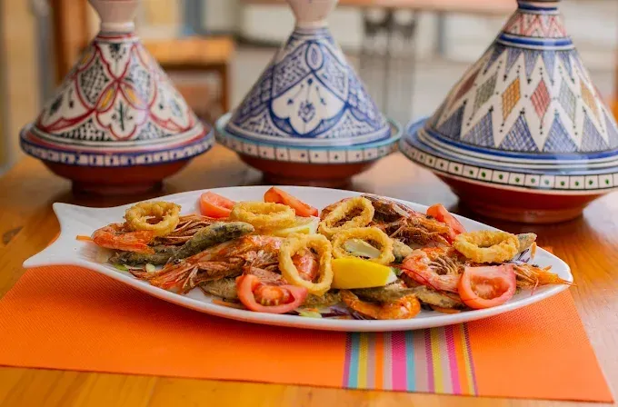 Plato decorativo con coloridos mariscos fritos sobre un mantel naranja, con ollas tagine marroquíes al fondo.