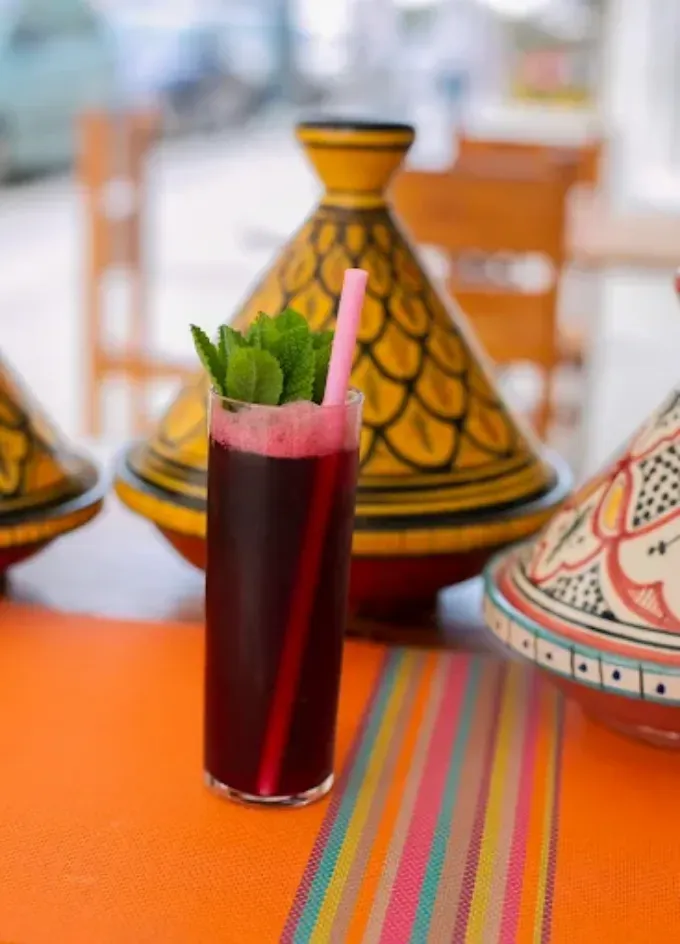 Bebida roja alta con menta y pajita rosa sobre una mesa de color naranja brillante junto a tagines marroquíes estampados.