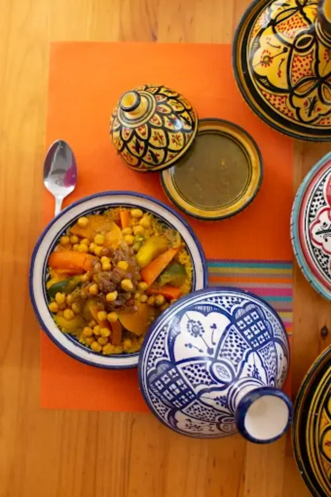 Platos coloridos al estilo marroquí con guisos y tapas estampadas sobre una mesa de madera, servidos sobre un mantel naranja.