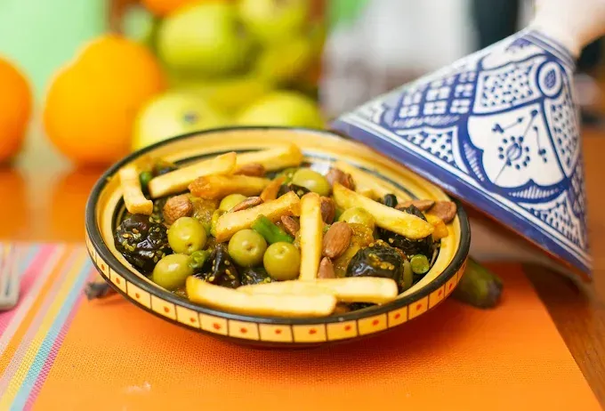 Bol decorativo con aceitunas y palitos de pan sobre una mesa naranja con tetera marroquí estampada.