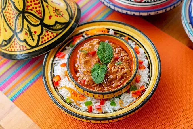 Un tazón de curry rojo en un plato colorido con estampado, acompañado de arroz y adornado con menta sobre un paño naranja.