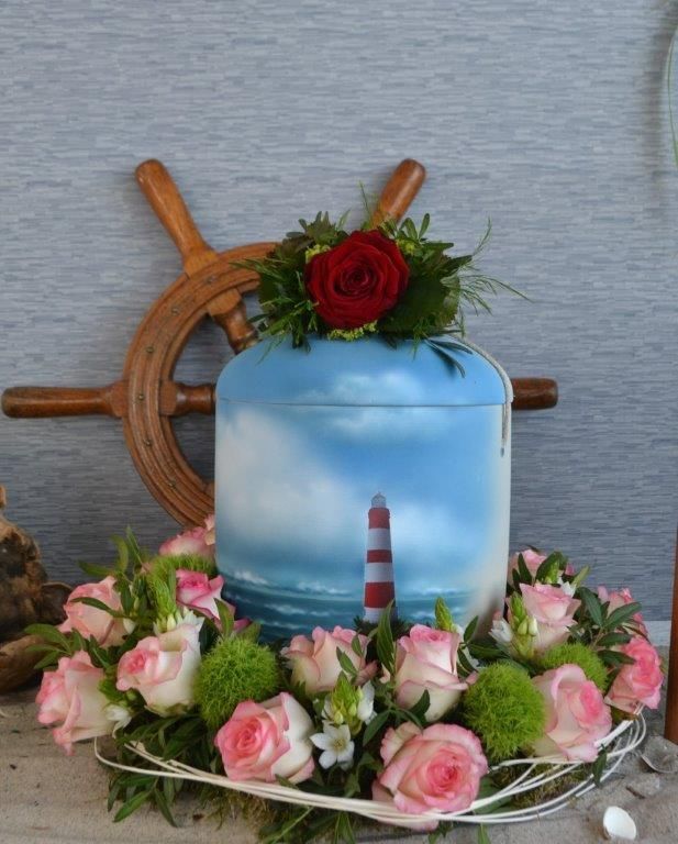 Eine blaue Urne mit einem Leuchtturm darauf ist von rosa Rosen umgeben