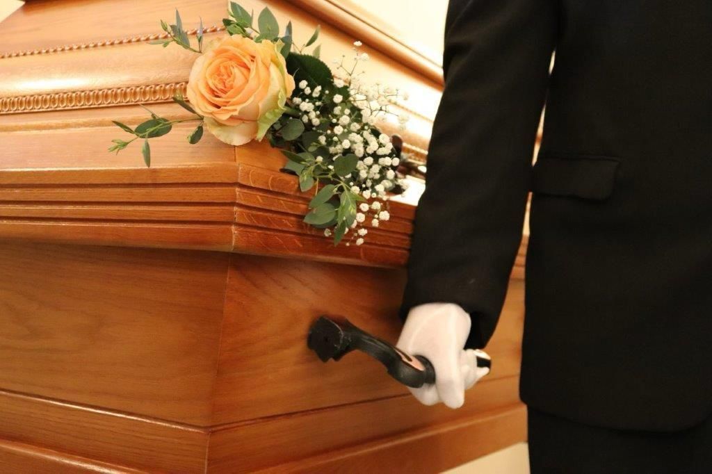 Ein Mann im Anzug hält einen Sarg mit Blumen darauf.