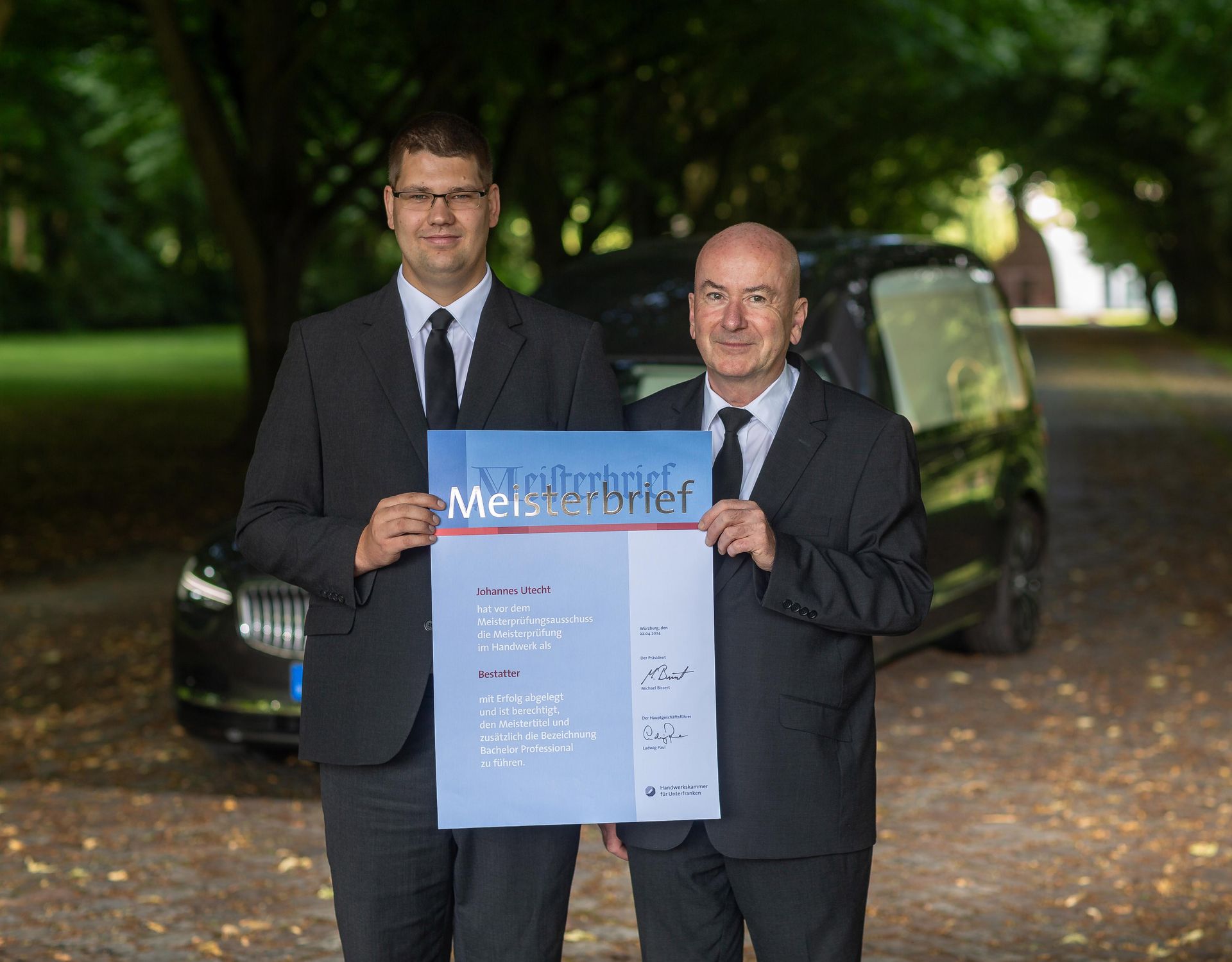 Zwei Männer in Anzug und Krawatte halten ein Schild vor einem Auto.