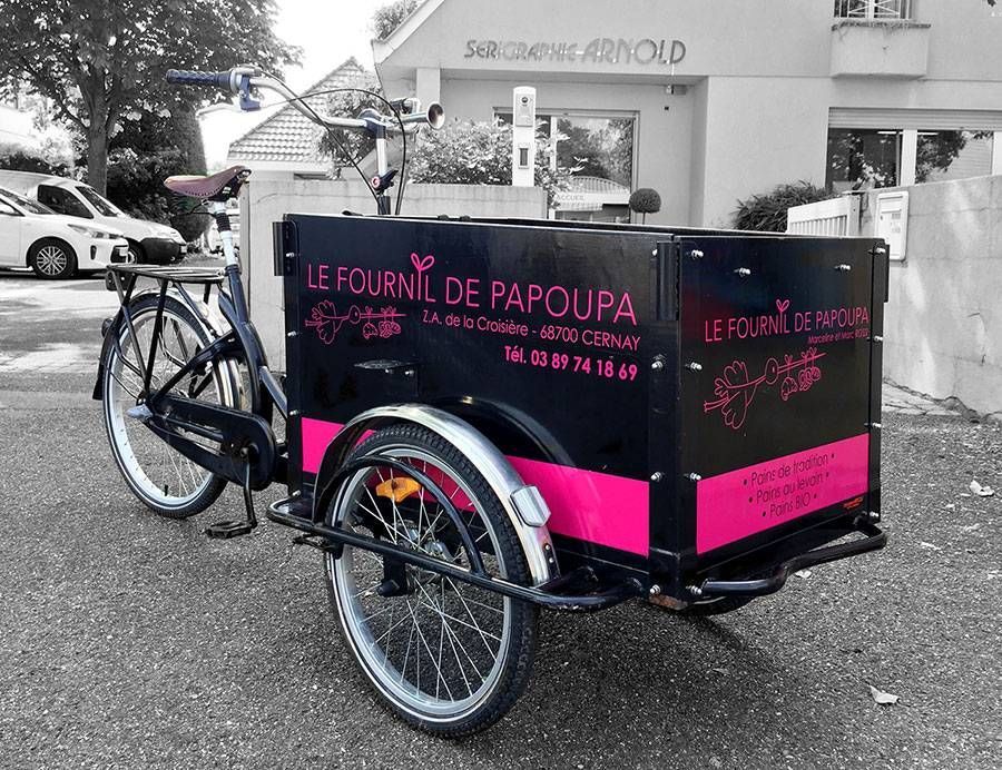 Triporteur cargo noir et rose stationné à l’extérieur, avec une signalétique en français et des enjoliveurs blancs