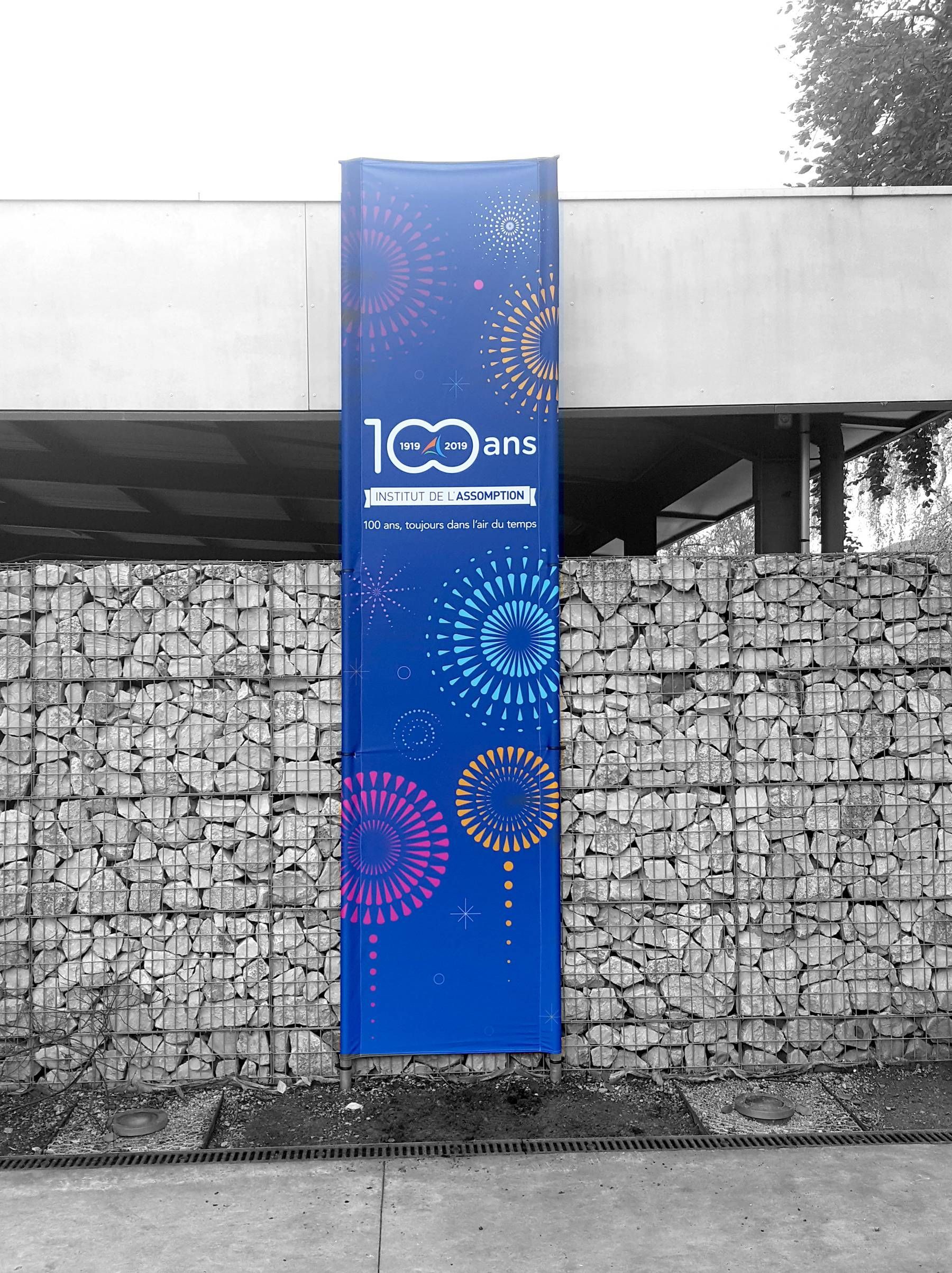 Bannière verticale bleue « 100 años » sur un mur en pierre à l’extérieur, ornée de motifs circulaires colorés.