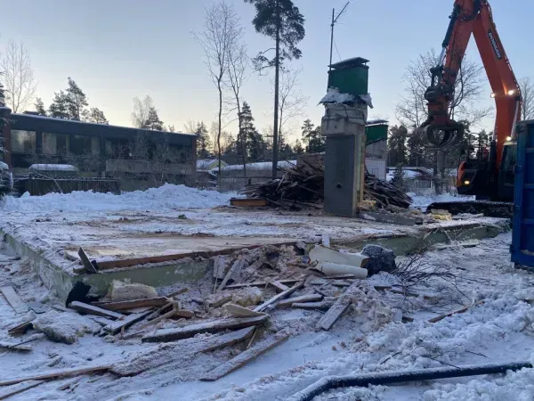 Purkutyömaa, jossa on raunioita, kaivinkone ja purettu rakennus lumisessa maisemassa hämärässä