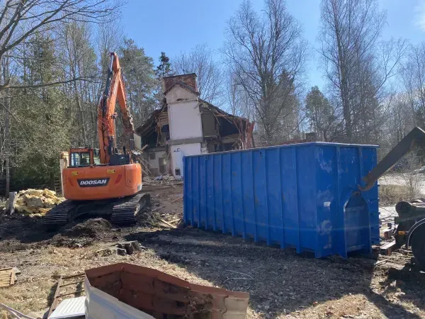 Rakennustyömaa, jossa oranssi kaivinkone sinisen roskalavan ja osittain puretun talon vieressä