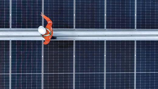 Ouvrier en tenue de sécurité orange sur un toit de panneaux solaires, inspectant ou nettoyant les panneaux au-dessus d'une passerelle centrale