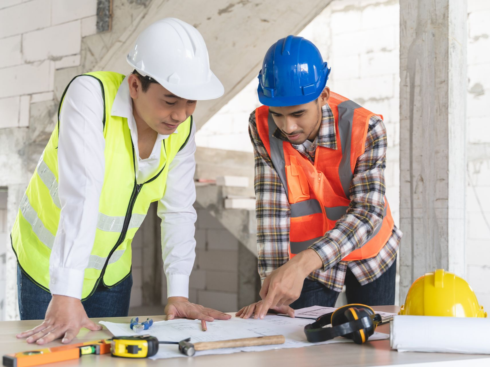 Deux ouvriers du bâtiment, casqués, examinent des plans sur un chantier de rénovation.
