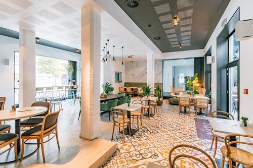 Intérieur du restaurant avec tables et chaises, sol carrelé décoratif, colonnes blanches et grandes fenêtres.