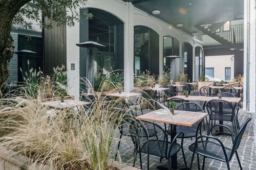Terrasse extérieure de restaurant avec tables, chaises et plantes ; architecture moderne avec fenêtres cintrées.