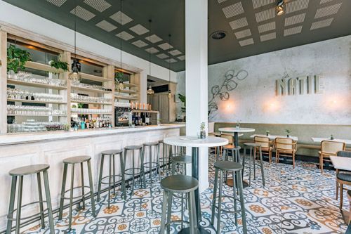 Intérieur d'un bar avec un comptoir et des tabourets blancs, un carrelage à motifs au sol et un plafond gris.