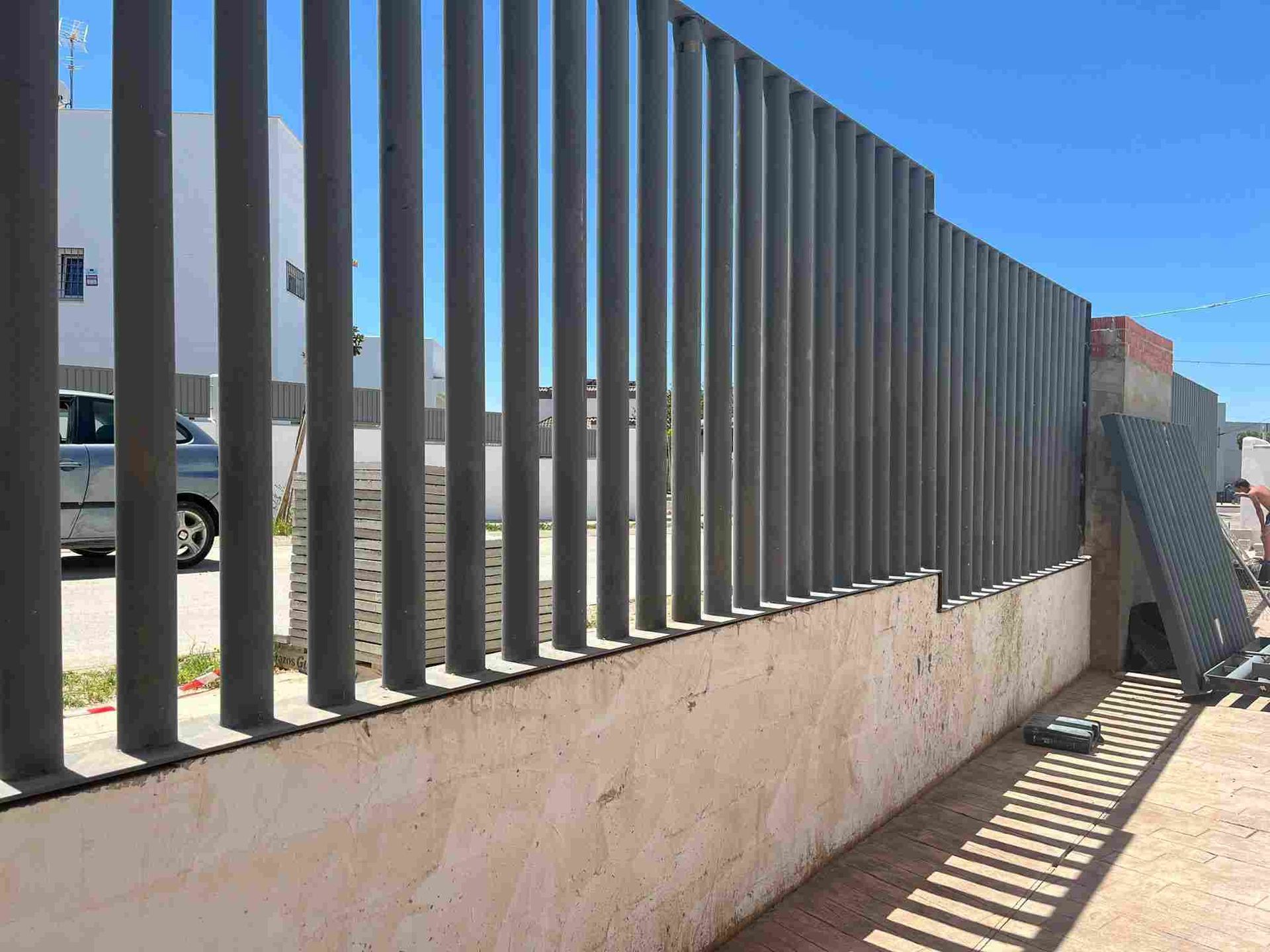 Valla de metal gris con barrotes verticales, base de hormigón y cielo azul.