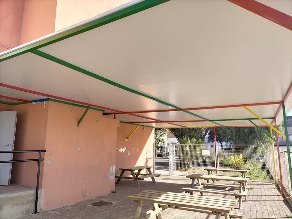 Zona de estar al aire libre cubierta con mesas de picnic. Ribete rojo, verde y amarillo. Edificio rosa.